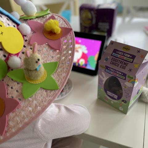Foreground: over the shoulder shot of a child wearing a hand made Easter bonnet with decorative bunny, chick and flowers. Child’s elbow on a table. In background on table: A “free from” Easter Egg box, a tablet playing a cartoon, and further away, possibly Easter Egg boxes blurred.