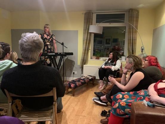 A living room in a house with people sitting on sofas and chairs. A blonde woman with a piano and a microphone is standing at the front performing.