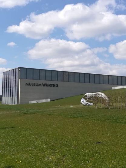 Gebäude Museum Würth2. Grüne Wiese mit einem Kunstwerk und blauer Himmel mit Wolken.