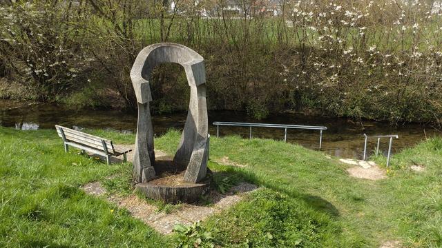 Ein aus einem Baum geschnitztes Kunstwerk, daneben eine Holzbank und dahinter ein Bach mit einem Kneippbad-Geländer im Wasser. Hinter dem Bach Büsche und dahinter  sind die ersten Häuser eines Dorfes erkennbar.
