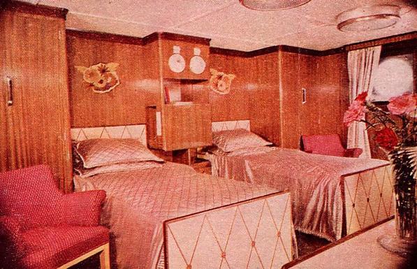 Color photo of a first class stateroom aboard the MV Britannic, 1950. There are two beds with white shiny quilted comforters. wardrobes and walls covered in the same reddish-brown paneling, and ugly abstract flower illustrations over the beds. A porthole is visible on the wall to the right.