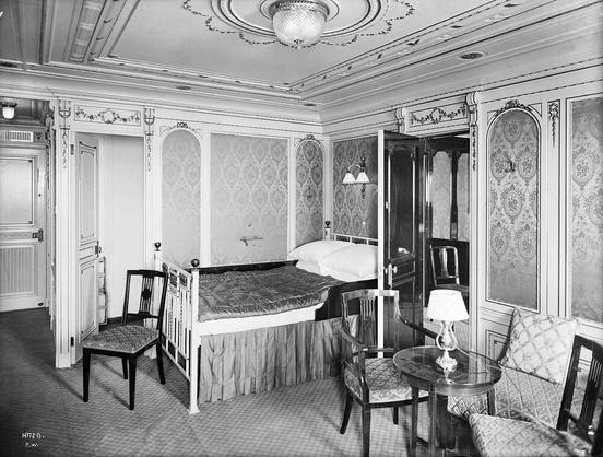 Black and white photo of the bedroom of suite B-56 aboard the Titanic, April 1912. There is a bed with satin comforter, silk wall coverings with white paneled edging, and a small round table with matching chairs. A door is open to what appears to be a walk-in closet (probably not as deep as it looks).
