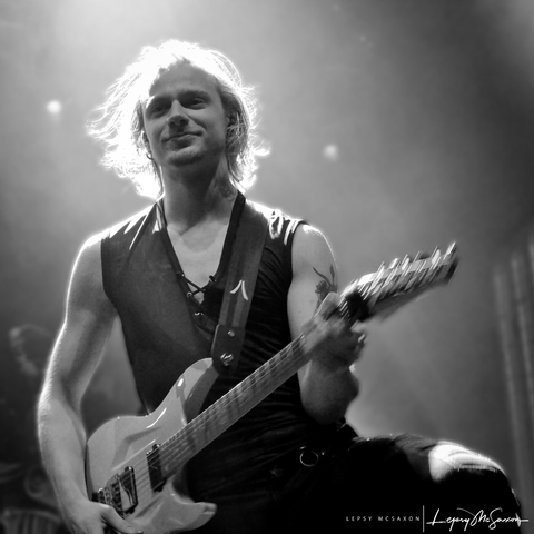 Ein schwarz-weiß Foto. Tim Hansen von Induction spielt grinsend Gitarre. Sein blondes Haar ist von hinten angestrahlt.

A black and white photo. Tim Hansen of Induction is grinning and playing guitar.