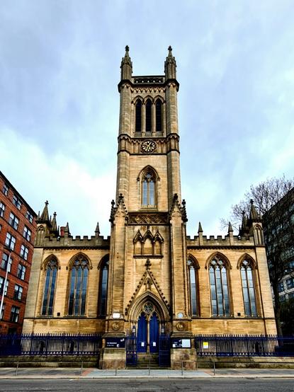 A low, two storey Gothic style church made of blonde sandstone with a tall clock tower in the middle.