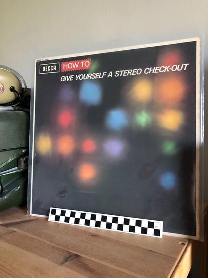 A 12-inch LP record sitting on a shelf next to a pair of vintage 1970s KOSS stereo headphones (made of beige plastic and metal) and an EMI dictaphone audio recorder (a green metal box). The LP record's cover is a blurry photograph of an abstract pattern of coloured lights on a black background shot through gauze to make the lights look fuzzy. On the shelf in front of the LP record is a measuring stick marked in centimeters for scale.