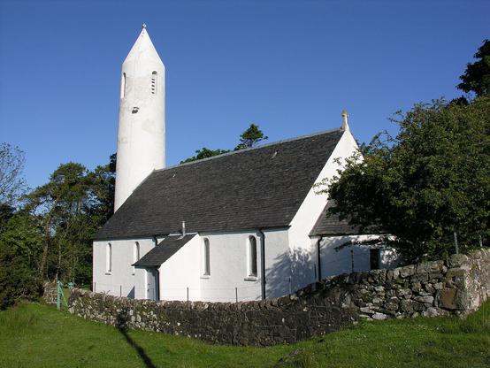 Kilmore Church at Dervaig. The image shows a rear quarter view of a white church beyond a grey stone wall. At the far end of the church is a white circular pencil-like tower. The sky is blue.