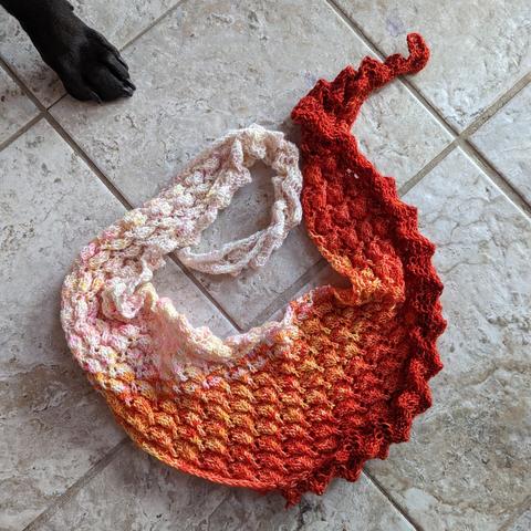 A small unblocked knitted shawl fading from white with pink and yellow speckles to a deeper tonal red. A dog's paw is visible in the upper left. Because it is unblocked, the shawl is heavily textured and the "back" side is filled with little 3d cones.