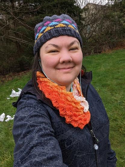 Terri is wearing her February Feather Shawl "backwards" so that the conical texture is pointed out. She's also wearing a knitted colourwork "Triangle Hat". She is standing in a mostly green backyard but there is some snow visible behind her.