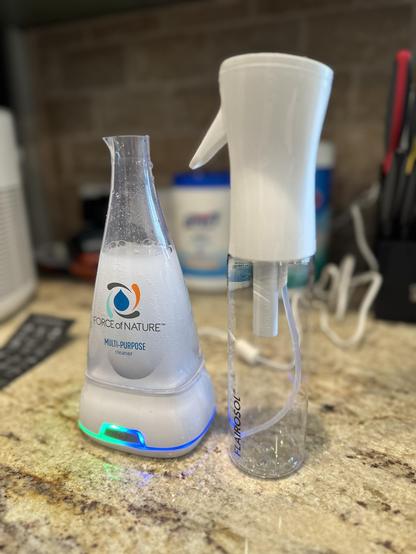 A Force of nature beaker in the process of creating HOCl solution (left) next to a clear empty misting spray bottle with a white lid (right)