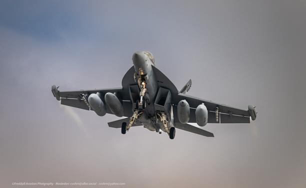 a all grey military aircraft is seen from the lower slight right on final. It has twin tails, two pods ont he wingtips, two fuel tanks and two tapered jamming pods underneath the wings. its gear is down and there are pronounces vapor trails coming from the wingtip pods that trail behind the aircraft. it is set against a white cloud background that is mostly opaque with a hint o blue in the upper left corner