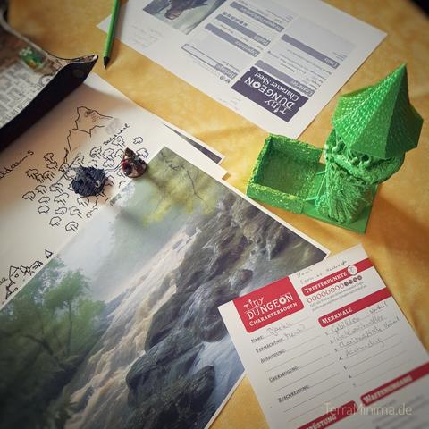 Draufansicht auf einen Tisch. Linksseitig quer über Bild liegen mehrere Papierblätter mit handgezeichneter Fantasy Map und Stimmunbsbild eines reißenden Flußes mit Bäumen an Flussufer. Auf den Papieren stehen Miniaturfiguren. Rechts oben und unten im Bild liegen je ein ausgefülltes Charakterblatt von Tiny Dungeon. Rechts mittig steht ein 3d gedruckter metallisch schimmernder Würfelturm mit spitzem runden Dach.
