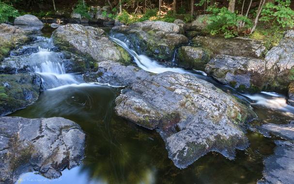 Rocks that have been smoothed by running water have a waterfall running smoothly white on either side and between them. The banks on either side have some green brush and small tree trunks visible. At the bottom of the fall are very smooth pools of dark water.
