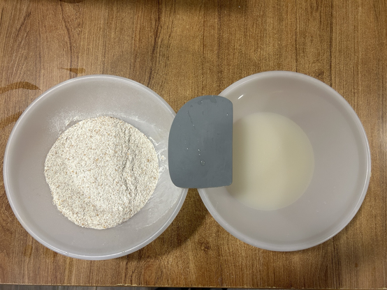 Closeup of two plastic mixing bowls sitting side by side, with my dough scraper resting on their rims. In the left bowl is the flour, in the right there's the water mixed with the yeast.