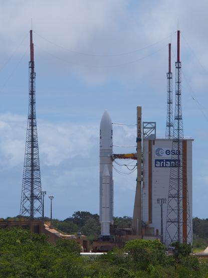 A rocket on a launch pad. Green trees are near the base, and the sky is blue with some clouds. The building next to the rocket is labelled "esa ariane" for the European Space Agency's Ariane rocket which will launch JUICE.