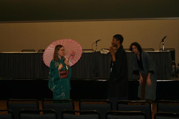 Three actors perform a Japanese inspired play in a hotel conference center panel room during an anime convention. Each wears garb inspired by Japanese historic culture. One is holding an umbrella and looking shocked at the story told by the other while the third is hiding to spy and looking outraged.