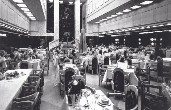 Black and white photo of the first class dining saloon aboard the SS Ile de France, 1927. The room is a vast open space, three decks high, with no visible columns except at the front (flanking a wall decorated with metal bas-relief). There are numerous tables all draped with white cloths, fine china, and stylish chairs that have a vaguely Art Deco style. The room is like a very large restaurant with a unified visual/design theme.