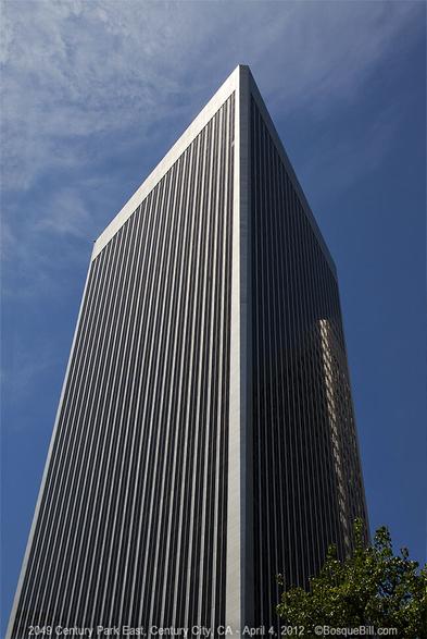 Photo shows two sides of a tall slender skyscraper. This modernist, minimalist building features uninterrupted, slim vertical lines between the windows on the sides with only a larger margin at the corners and top. A blue sky with thin clouds.
©BosqueBill.com