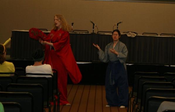 Two actors perform a Japanese inspired play in hotel conference panel room during an anime convention, each wearing garb inspired by historic Japanese fashion and playing a bickering wife (who is looking at a prop fan standing in for a mirror) and her husband.