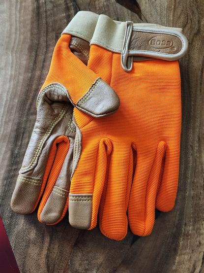 A pair of brand new, unworn, glowy orange gardening gloves laid on a table, palms together. They're clean and pristine, waiting to be put to work.