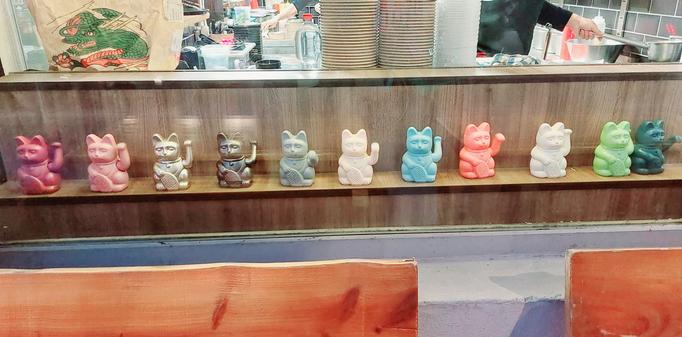 Schaufenster eines Schnellimbisses, in dem 11 Winkekatzen in verschiedenen Farben in einer Reihe stehen.
