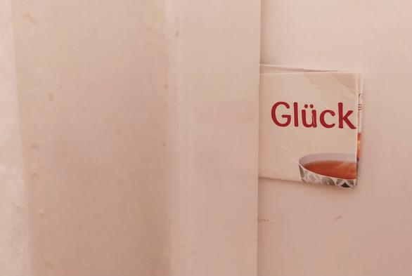 Pappe, die zwischen Tür und Rahmen steckt. Darauf das Wort Glück und ein Stück einer Teetasse sichtbar.