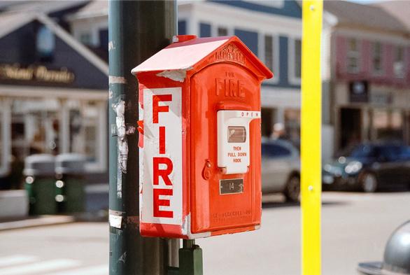 A color photograph of a bright red fire alarm call box, marked  as number 14, mounted to a vertical dark green metal lamp post on the side of the road. A slightly blurred fluorescent yellow strip  punted to a signpost rises through the whole frame on the fight, and the. Businesses in the background on the opposite side of the street are slightly defocused.