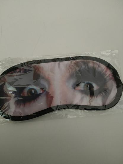 A plastic wrapped sleep mask featuring Reuben Kaye's eyes in full drag makeup including excessive amounts of tarantula-like false eyelashes. There is a thick black edging to keep Reuben's eyes from fraying, which helps amplify the overall visual effect.