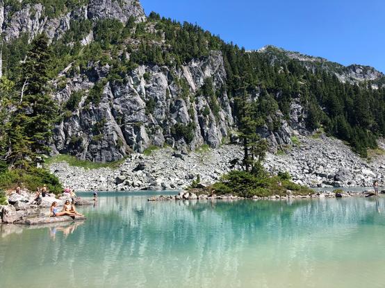 A mountain lake with people dotting the shoreline and steep treed cliffs in the background