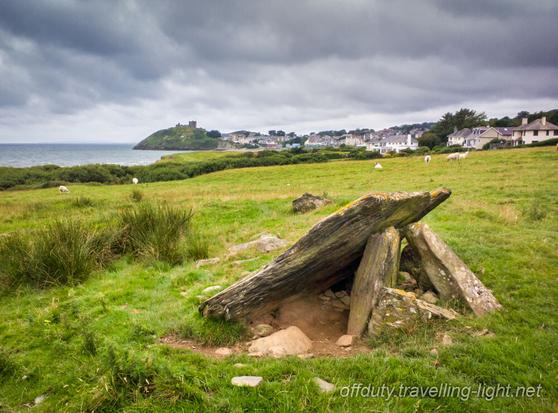 A collapsed prehistoric burial chamber in a field, with a castle on a coastal crag in the distance.