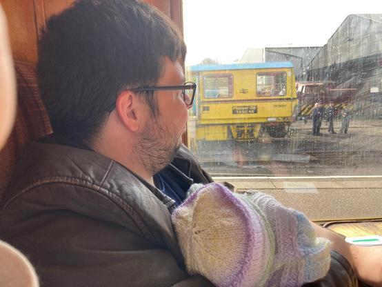 Man in leather jacket looking out of a railway carriage window, holding a newborn baby.