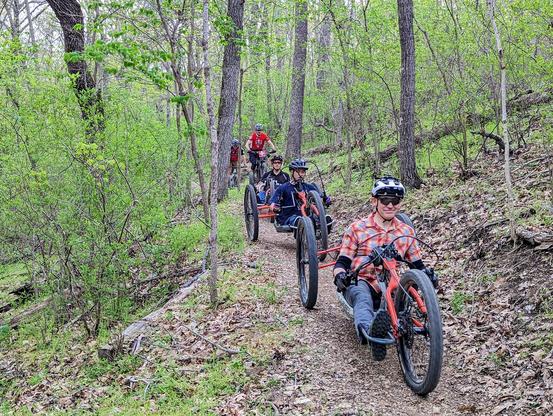 Adaptive Mountain bikers on a dirt trail in the forest being followed by trail patrol on regular mountain bikes. Adaptive Mountain Bikes are modified for wheelchair users to ride in a seated or prone position. Pictured here are seated riders on bikes with 2 wheels in the back and one in the front. They are hand powered with electric assistance.