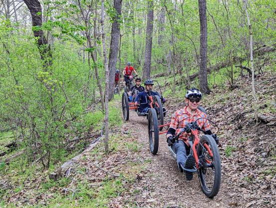 Adaptive Mountain bikers on a dirt trail in the forest being followed by trail patrol on regular mountain bikes. Adaptive Mountain Bikes are modified for wheelchair users to ride in a seated or prone position. Pictured here are seated riders on bikes with 2 wheels in the back and one in the front. They are hand powered with electric assistance.