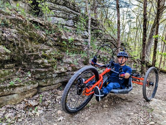 A young adaptive mountain biker riding a 3-wheeled bike on rocky terrain next to a small rock bluff