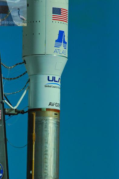 a photograph of the section joining the lower stage of the Atlas 5 rocket with its 5 meter diameter fairing, as well as several umbilical links stretching from the rocket to the Mobile Launch Platform’s tower
