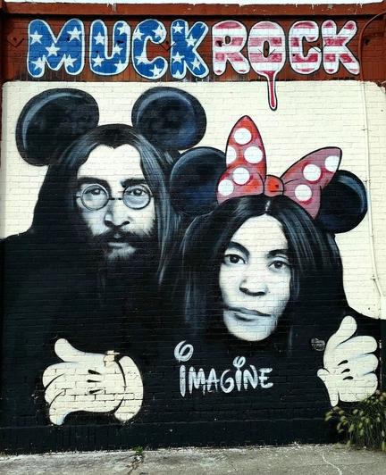 Streetartwall. On a red brick wall is sprayed / painted a portrait of the John Lennon and Yoko Ono. 
The background is designed in white, the figures in black and white. Both wear long black hair, Lennon a full beard and nickel glasses. On their heads they have black Mickey Mouse ears, and Yoko Ono has additionally a red and white Minnie Mouse hair bow. They hug each other with MickyMouse gloves.
The two are looking at us.
The name of the artist MuckRock with the colors and American flag elements is above it and the word "Imagine" completes this tribute to the two.