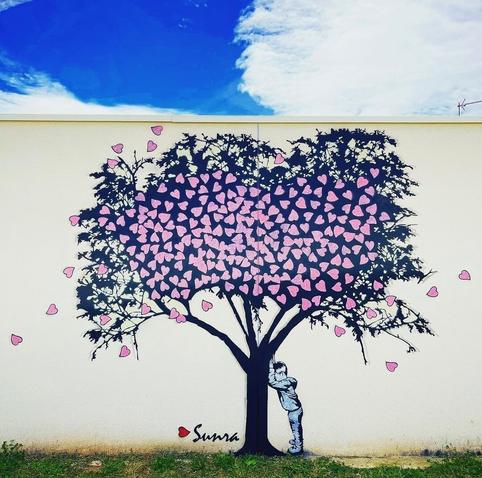 Streetartwall. On a white wall of a kindergarten is sprayed / painted the charming mural of a large tamaris tree full of pink hearts. A little boy leans his head against the tree, whose leaves are made of hearts and seems to count to 10. Everything must be hidden...
Some hearts blowing away in the wind....
