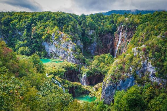 A color view looking down into a canyon topped by a thick green forest. There are two emerald colored lakes separated by waterfalls. On a sheer canyon wall a thin tall waterfall descends over an orange colored cliff.