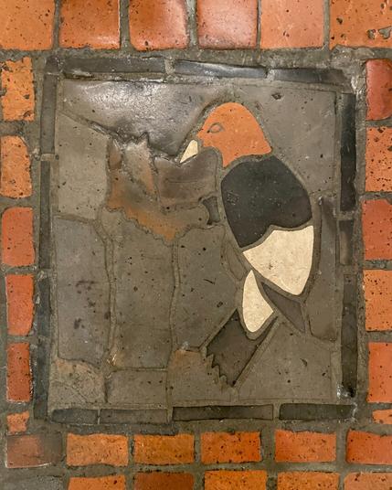 small tile mosaic depecting a red-headed woopecker in side profile, on grey background with black rectangular frame on red tile floor
