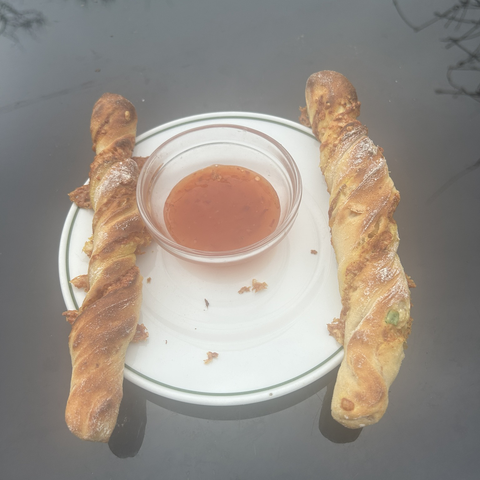 Two cheese sticks, on a plate, with a little bowl of sweet chilly sauce in between. The plate is sitting on my outside table and in natural light the cheese sticks have a soft golden sheen. Also visible: a bit of the added fruit peel (one of the green pieces) is peeping out through the outer layer of the bread.