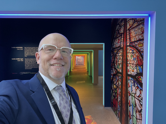 Selfie: a bald man with white glasses and a close-cropped beard wearing a blue suit, white shirt, and silver tie in front of a bunch of rectangles in different colors.