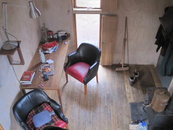 the interior of the both - a wood stove, a modern leather chair, a writer's desk, the clutter of travel: books, walking boots, a hot water bottle