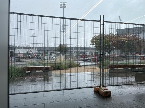 A view out a window. Cyclone fencing in foreground and workers running for cover behind it.  It’s raining hard and it’s grey.