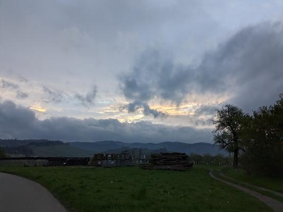 Blick in Richtung Sonnenaufgang. Im Vordergrund ist eine Wiese auf der ein Landwirt sein Brennholz gestapelt hat. In der Bildmitte sind die Gebirgszüge der Ausläufer des Schwarzwaldes zu sehen und am Himmel sieht man dramatische Wolken mit aufgehender Sonne im Hintergrund