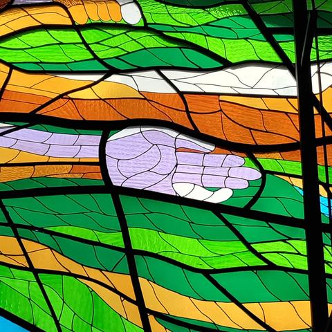 Detalle de un vitral emplomado con piezas de color verde, amarillo, naranja, gris y azul. Una mano humana se destaca en lo que parece una corriente o un viento.