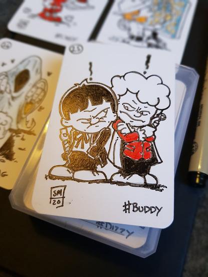 A photo of an ink doodle on a blank playing card. Drawn in a Calvin and hobbes styles. The 2nd and 3rd Doctors are standing facing slightly away from each other. Both heads turned to look at each other, and both look none to please to be spending time together. 2 has his hand stuck in his pockets with a grumpy face, and 3 has his arms crossed, sonic screwdriver in hand and has an annoyed expression.
