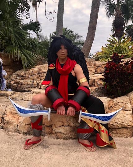 Dark Pit cosplay from Kid Icarus by MelPool. Black spiked wig, black wings, red scarf, red and gold cap on right shoulder, black tunic, red and black leather forearm guards, blue and silver bow, gray and red leather shin armor, brown and red sandals.