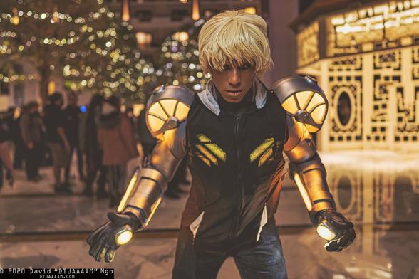 Genos cosplay from One-Punch Man by MelPool. Blonde wig, sclera eyes (edited by photographer), silver shoulder armor with yellow lighting, black transparent hoodie with chest lighting underneath, silver forearm armor with yellow lighting, yellow palm lighting.
