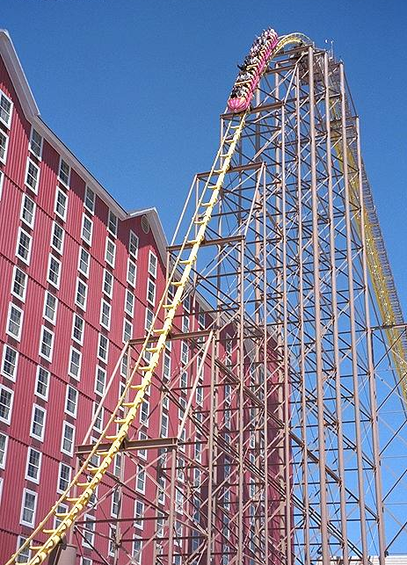 A picture of a train car descending the tallest hill on Desperado, shot from the ground.  The track is painted bright yellow with tan supports.  The train car is painted pink with yellow accents.  Directly behind the roller coaster is Buffalo Bill's hotel, red with white trim around the windows.  These guest rooms had a very up close look at the roller coaster when operating.