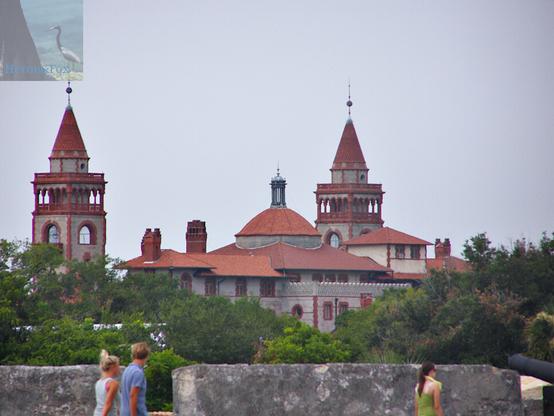 A photograph of a building in the distance rising over some trees. The building is large and ornate, made of stone with terracotta tiles. There are two towers topped with cones and a central dome.