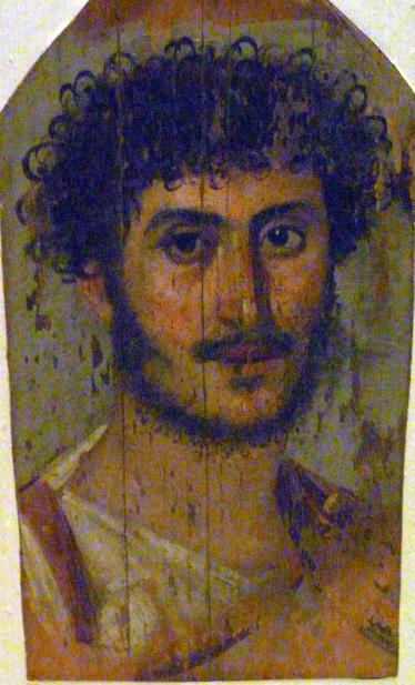 Portrait of a young Roman created using encaustic painting techniques. He engages the viewer directly, looking calm and composed. He sports a head of dark, curly hair, and a sparse if equally curly beard, and moustache.